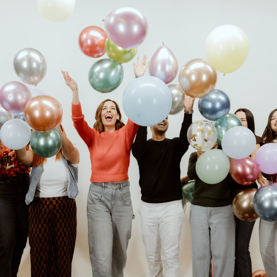 A group of people standing and smiling while tossing colorful balloons into the air against a plain background.