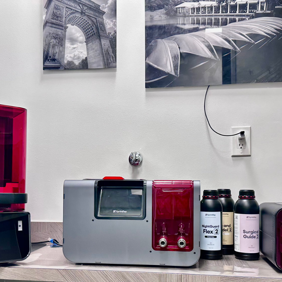 A 3D printer and resin bottles are on a counter beneath black and white photos on a wall. The setup appears to be in a professional or laboratory environment.