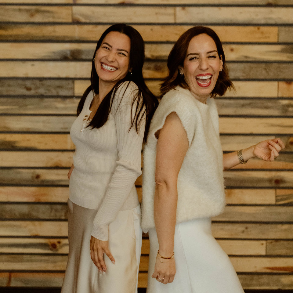 Two women standing back to back and smiling, with a wooden plank wall in the background.