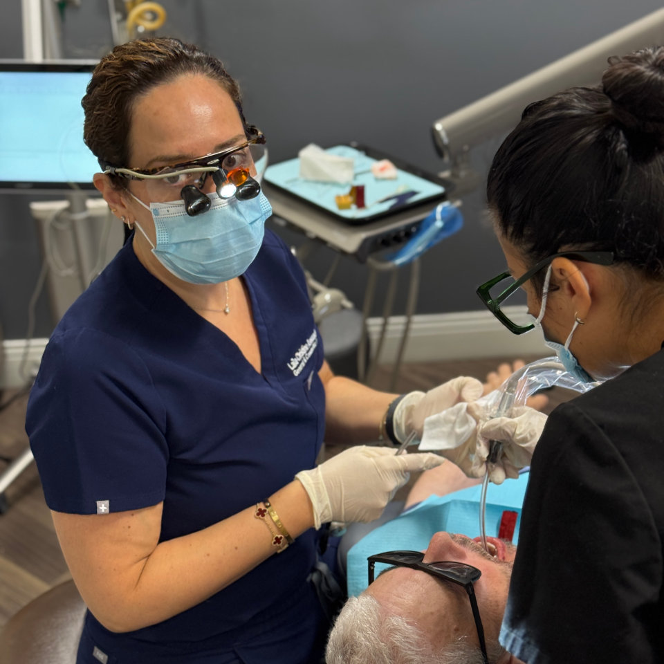 Two dental professionals in scrubs and masks perform a dental procedure on a patient lying in a dental chair, using medical instruments and wearing protective eyewear.