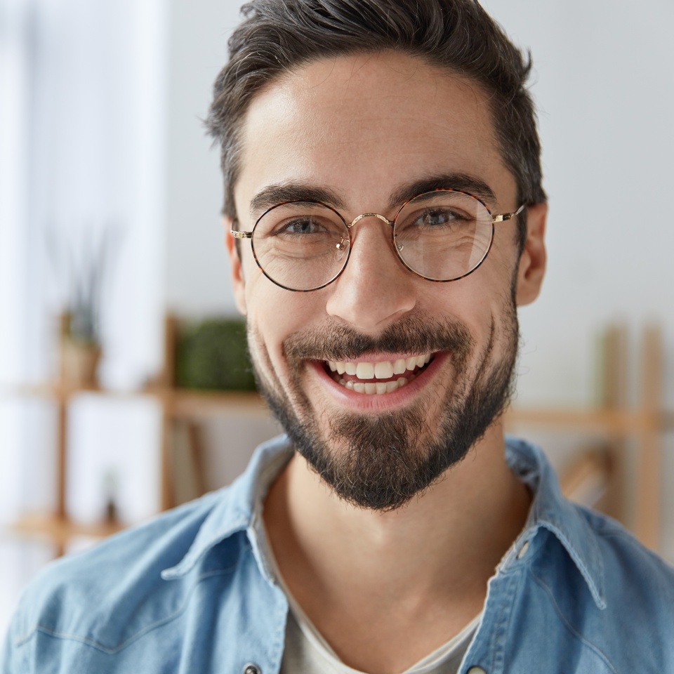 Close up shot of cheerful satisfied attractive male with stubble, has broad smile, wears round spectacles, rejoices success at work, stands against cozy interior. Fashionable designer glad be praised