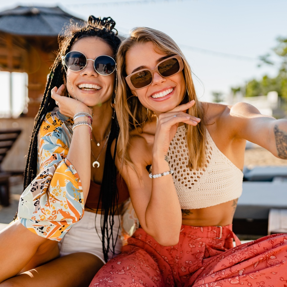 stylish smiling beautiful women relaxing having fun on beach in summer fashion trend outfit, colorful apparel boho style, tropical vacation wearing sunglasses, using smartphone taking selfie picture