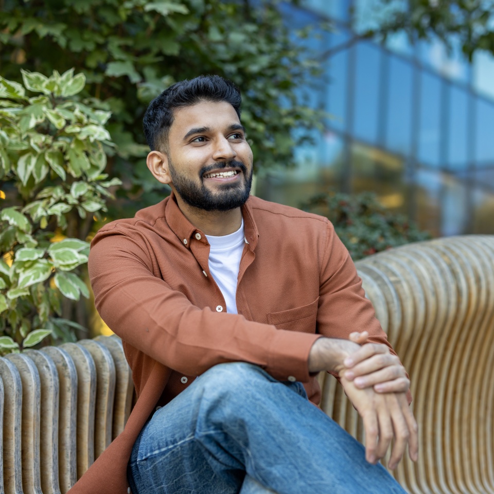 A smiling man in a casual outfit sits on a wooden bench, enjoying a sunny day outdoors with a modern building backdrop.