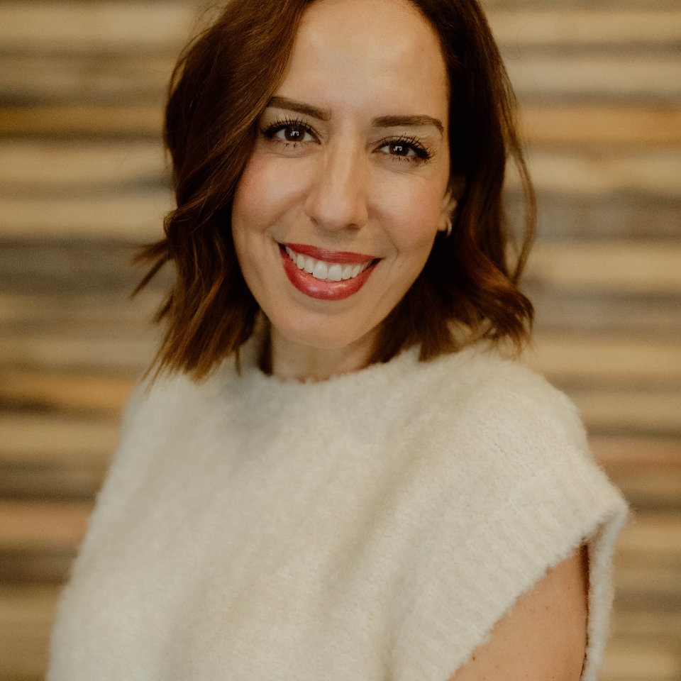 Woman with shoulder-length brown hair and a white sleeveless top smiles in front of a blurred striped wooden background.
