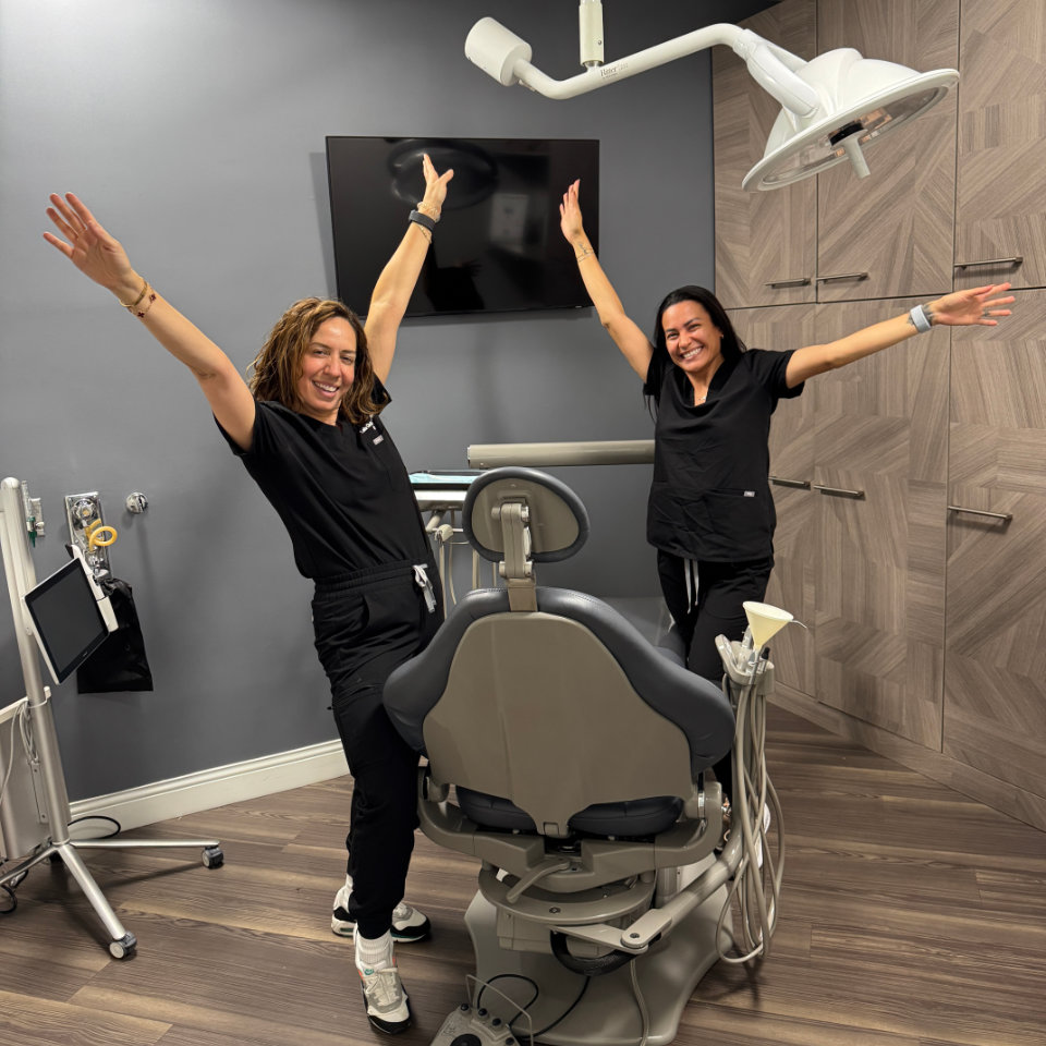 Two women in black scrubs stand with arms raised, smiling, next to a dental chair in a modern dental office.