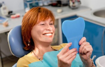 Smiling senior woman with new dental implants sitting in the dental office and looking at the mirror
